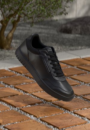 Sort Tommy Hilfiger-sneaker med snørebånd, svævende over træfliser og grå småsten, med en lille grøn plante i baggrunden.