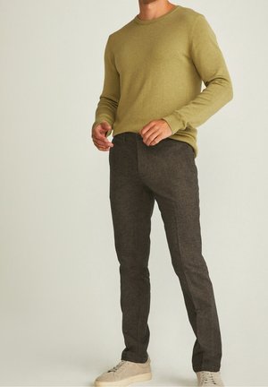 Mann trägt einen olivgrünen Pullover mit langen Ärmeln, dunkelgraue eng anliegende Hose und hellbeige Turnschuhe, steht mit den Händen nahe der Taille.
