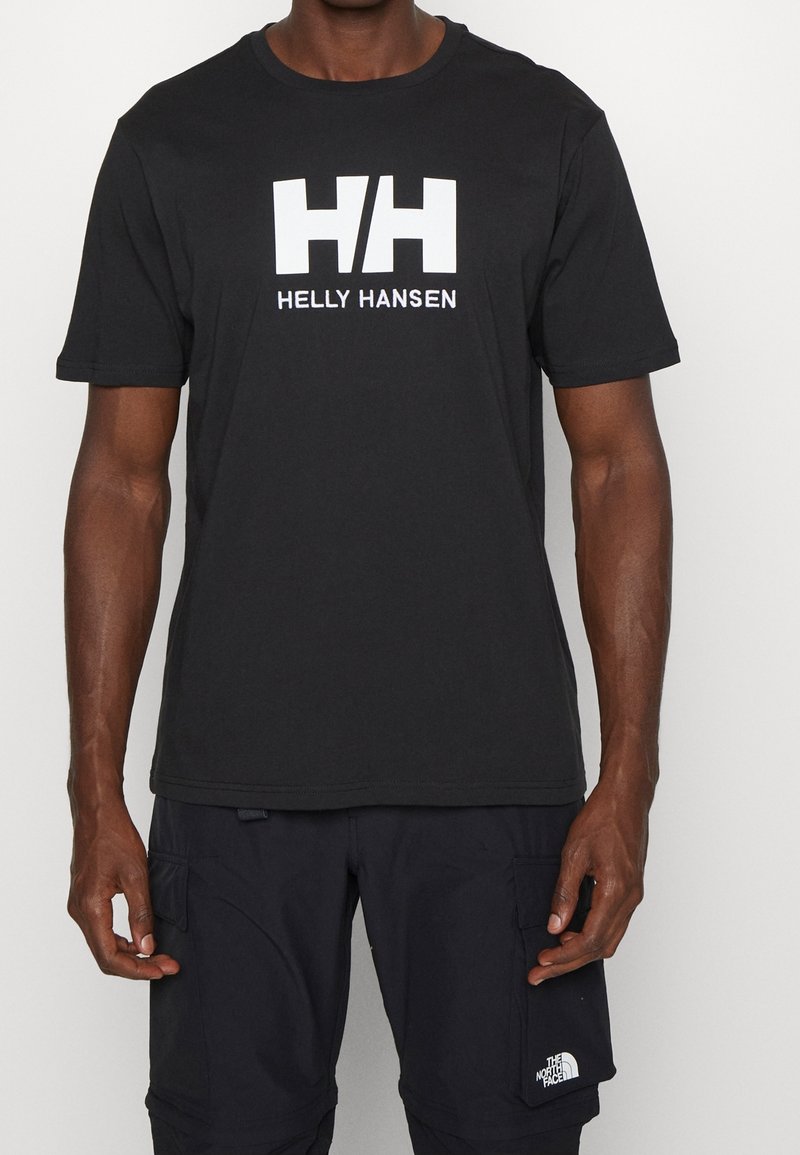 Homme portant un t-shirt noir Helly Hansen avec un logo blanc et un short cargo noir avec le logo The North Face sur la poche droite.