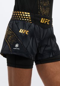 Črni UFC kratki hlače imajo zlati geometrijski vzorec na pasu in abstraktno obliko. Vključujejo logotip "UFC" in blagovno znamko na nogi.