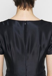 Zadní pohled na osobu, která má na sobě černé saténové šaty s viditelným malým zipem ve středu zad.