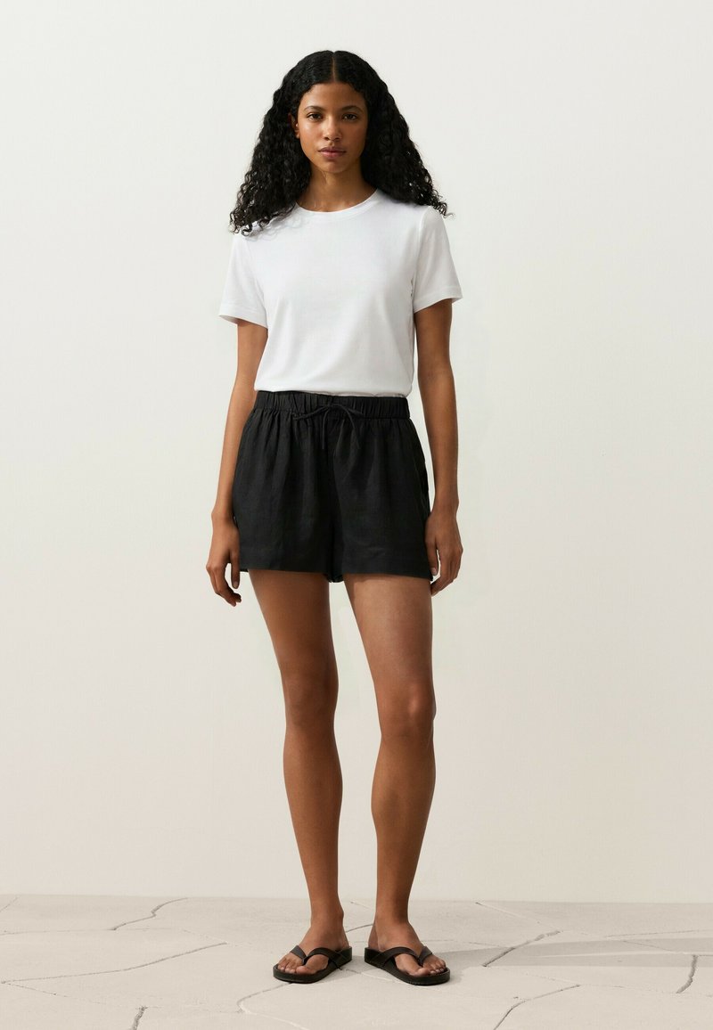 Femme aux cheveux bouclés portant une chemise blanche à manches courtes, un short noir à cordon de serrage et des sandales noires, debout sur un sol carrelé.