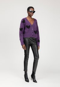 Donna con occhiali da sole neri, cardigan viola con cuori neri, pantaloni in pelle neri e stivali con tacco alto neri, in piedi su uno sfondo bianco.