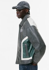 Giacca di pelle grigia con un design verde e bianco. Presenta tasche laterali e un colletto. Indossata con un cappellino grigio.