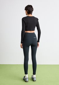 Kvinna som står på grön golv iklädd svart crop-top, mörkgröna Columbia leggings, vita strumpor och silvergrå sneakers, vänder ryggen mot kameran.