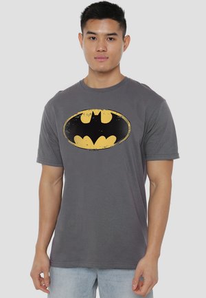 Young man wearing a gray t-shirt with a distressed yellow and black Batman logo, standing against a plain light background.
