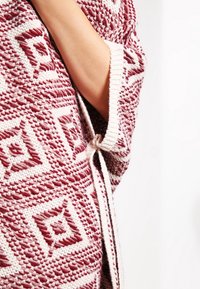 Strikket cardigan med et geometrisk rødt og creme mønster, der har lukning med løkke og åbne ærmer, som fremhæver teksturerede stofdetaljer.