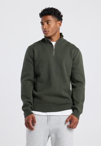 Dark green, zip-up sweater with a high neck, ribbed cuffs and hem. Made from textured fabric, worn over a white shirt and gray pants.