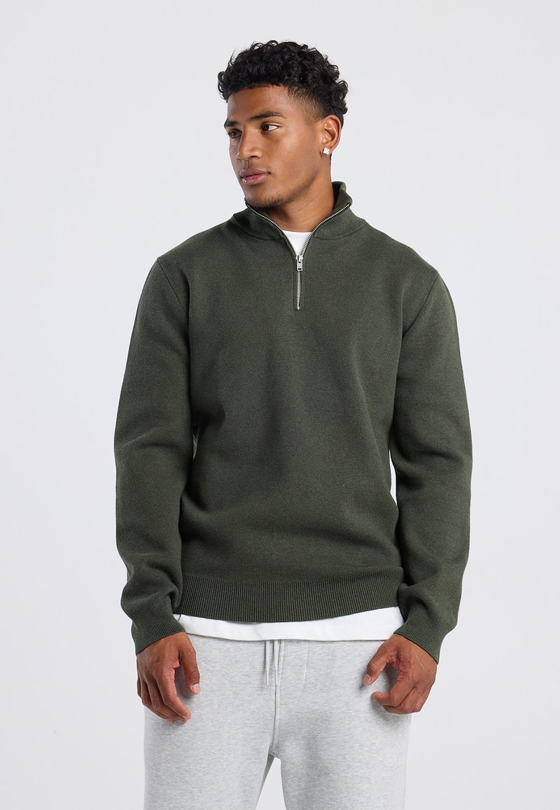 Dark green, zip-up sweater with a high neck, ribbed cuffs and hem. Made from textured fabric, worn over a white shirt and gray pants.