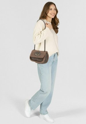 Jonge vrouw staand, gekleed in een crèmekleurige trui, lichtblauwe spijkerbroek, witte sneakers, en draagt een bruine schoudertas met een gestructureerd oppervlak en een kettingriem.