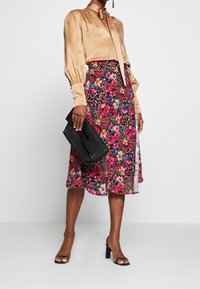 Blouse en soie avec des manches bouffantes en beige, jupe midi florale en rose vif et noir, et un sac à main noir. Des sandales à talons hauts complètent le look.