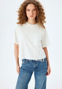 Camiseta blanca recortada con un bolsillo de encaje texturizado y detalles de flecos, combinada con jeans de mezclilla azul. Tejido suave, ajuste relajado.