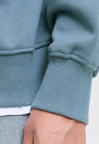 Közelkép egy személy csuklójáról, aki kék pulóvert visel bordázott mandzsettákkal és alatta fehér inget, világoskék farmerral párosítva.