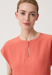 Korallefarbene Bluse aus strukturierter Stoffqualität, mit rundem Ausschnitt und Schlüssellochdetail vorne. Kurze Ärmel sorgen für eine lockere Passform.