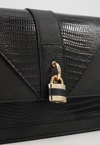 Czarna torebka o teksturowej powierzchni, z wzorem przypominającym skórę jaszczurki, złotymi akcentami i detalem zapięcia. Strukturalny design z eleganckim wykończeniem.