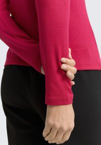 Person trägt ein rotes Langarmshirt und schwarze Hosen und hält den entgegengesetzten Arm hinter dem Rücken vor einem schlichten Hintergrund.