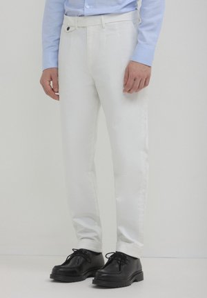 Homme portant un pantalon slim blanc, une chemise bleu clair et des chaussures en cuir noires à lacets, se tenant contre un fond clair et uni.