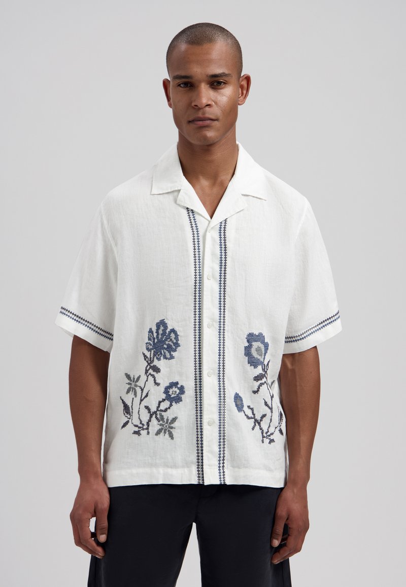 Homme portant une chemise blanche à manches courtes avec des broderies florales bleues et un pantalon noir, debout devant un fond uni.