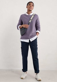 Läppäräinen tekstuuritakki violettina valkoisen kauluspaidan päällä, tummansiniset cargo-housut ja valkoiset tennarit. Istuva musta olkalaukku, jossa on kuvioitu hihna.