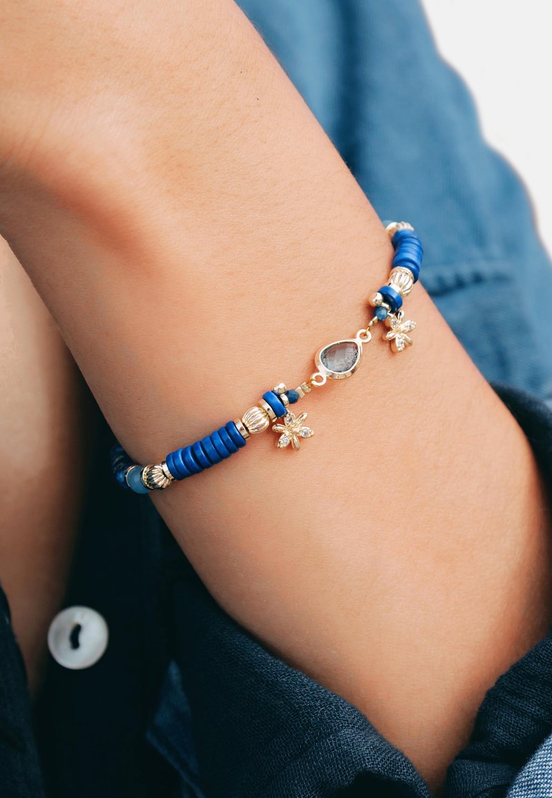 Bracelet en perles bleues avec des accents dorés, des détails en argent et des charms en forme de fleurs décoratifs. Texture lisse avec différentes formes de perles.