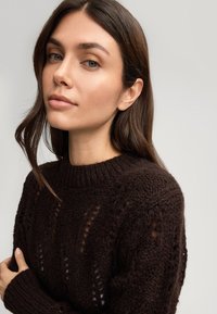 Ciemnobrązowy sweter dziany z prążkowanym dekoltem i slim-fit rękawami. Posiada otwarte wzory szycia oraz teksturowany, przytulny wygląd.