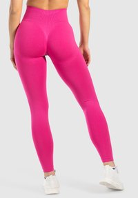 Korkeavyötäröiset vaaleanpunaiset legginsit, joissa on sileä, joustava pinta ja muotoileva suunnittelu, yhdistettynä valkoisiin lenkkareihin neutraalilla taustalla.