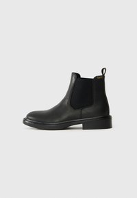 Botín Chelsea de cuero negro con paneles laterales elásticos, punta redondeada y una lengüeta. Textura suave con un tacón bajo apilado.