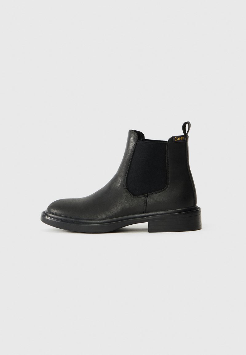 Botín Chelsea de cuero negro con paneles laterales elásticos, punta redondeada y una lengüeta. Textura suave con un tacón bajo apilado.
