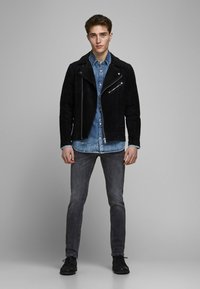 Schwarze Wildleder-Bikerjacke mit Reißverschlusstaschen, über einem hellblauen Jeanshemd getragen. Kombiniert mit grauen Jeans und schwarzen Schuhen.