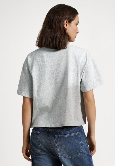 Žena s tmavými vlasmi po ramená, oblečená v voľnom sivom tričku s krátkym rukávom a modrých džínsoch, stojaca chrbtom k bielej stene.