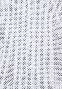 Bílá košile s tmavě modrými kruhovými vzory, s knoflíkovou légou vpředu a hladkou texturou. Výrazné detaily zahrnují malé rovnoměrně rozmístěné tečky.