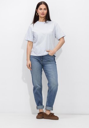 Femme aux longs cheveux foncés portant un t-shirt gris clair oversize, un jean bleu retroussé, des chaussures marron à lacets et des boucles d'oreilles créoles en or, debout contre un mur blanc.