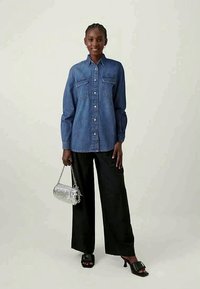 Chemise en denim à boutons avec deux poches poitrine, associée à un pantalon large noir. Le mannequin tient un sac à main argenté et porte des sandales noires à enfiler.
