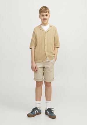 Chemise beige en tricot à manches courtes avec des boutons, t-shirt blanc en dessous, shorts beige clair et baskets teal et blanches avec des semelles marron.