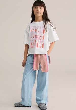 Balta oversized T-krekls ar rozā grafiku, kurā attēloti pārtikas produkti un teksts "BRUNCH", kopā ar gaiši zilām platām džinsa biksēm un rozā džemperu nošu lentu.