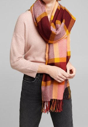 Personne portant un pull rose pâle et un jean foncé, tenant une grande écharpe à carreaux aux nuances de rouge, rose, orange et jaune avec des franges aux extrémités.