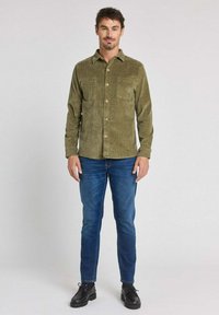 Chemise en velours côtelé vert olive avec fermeture à boutons et deux poches poitrine, associée à un jean denim bleu et des chaussures noires.