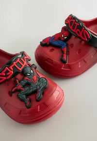 Zoccoli rossi in gomma con cinturino nero, dotati di design stampato di Spider-Man. Il testo sul cinturino recita "SPIDER." Fori di ventilazione sui lati.