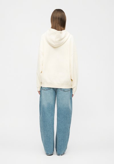 Personne aux cheveux bruns longs vue de dos, portant un sweat à capuche surdimensionné couleur crème et un jean large bleu clair, sur un fond uni.