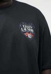 Μαύρο μπλουζάκι με λογότυπο σε σχήμα καρδιάς που γράφει "Lyle's Laundry, Making Fresh Look Easy, 150 Χρόνια Υπηρεσίας, 1874" στο αριστερό στήθος.