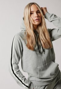 Sudadera gris hecha de tela suave, con un bolsillo frontal y cordones negros. Acentos de rayas verticales en blanco y negro en las mangas.