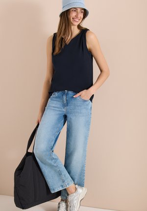 Femme portant un chapeau bob bleu clair, un haut noir sans manches, un jean bleu clair, des baskets argentées, tenant un grand sac cabas noir, souriant sur fond beige.
