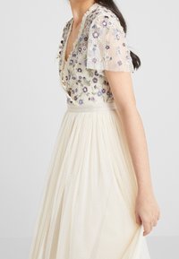 Cremfarbenes Kleid mit floraler Stickerei in Lila und Grün. Kurze Puffärmel, V-Ausschnitt und ein voller, fließender Rock mit Struktur.