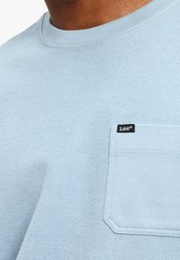 Närbild av en ljusblå texturerad skjorta med en bröstficka som har en liten svart Lee-märkesetikett.