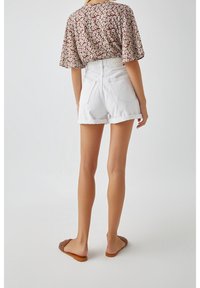 Femme portant un short en denim blanc à ourlet roulé, un chemisier marron à fleurs, et des sandales à brides tressées marron, debout devant un fond blanc uni.