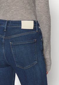 Dark blue denim jeans with a smooth texture, featuring a white leather tag at the back, and subtle stitching details on pockets.