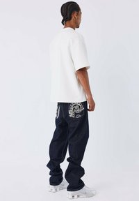 Witte oversized shirt met korte mouwen, donkerblauwe jeans met paisley borduursel op de zakken, gecombineerd met witte sportschoenen.