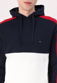 Navy blue and white hooded sweatshirt featuring red shoulder accents, a front pocket, and a small logo on the chest. Soft fabric texture.