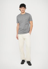 Camiseta de manga corta de punto gris con un sutil patrón de logo, combinada con pantalones blancos y zapatos negros; diseño minimalista y tejido ligero.