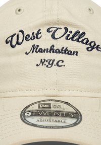Beige bomullskaps med justerbar rem, med marineblå broderte tekster: "West Village," "Manhattan," og "N.Y.C." på framsiden.